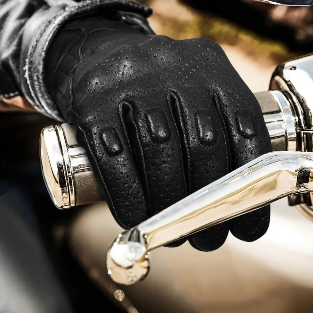 Black leather perforated motorcycle gloves with knuckle protection, worn on hand while gripping a motorcycle handlebar, showcasing touchscreen functionality